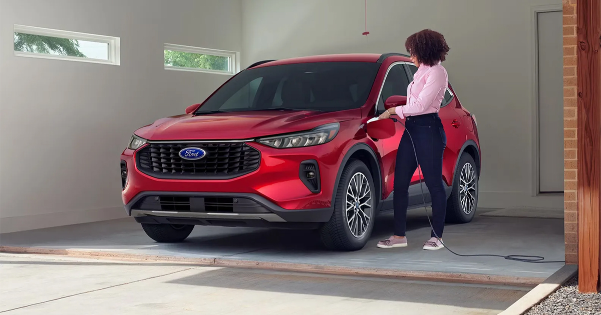 Red Ford Escape SUV being charged in a garage, with a person standing nearby plugging in the charging cable