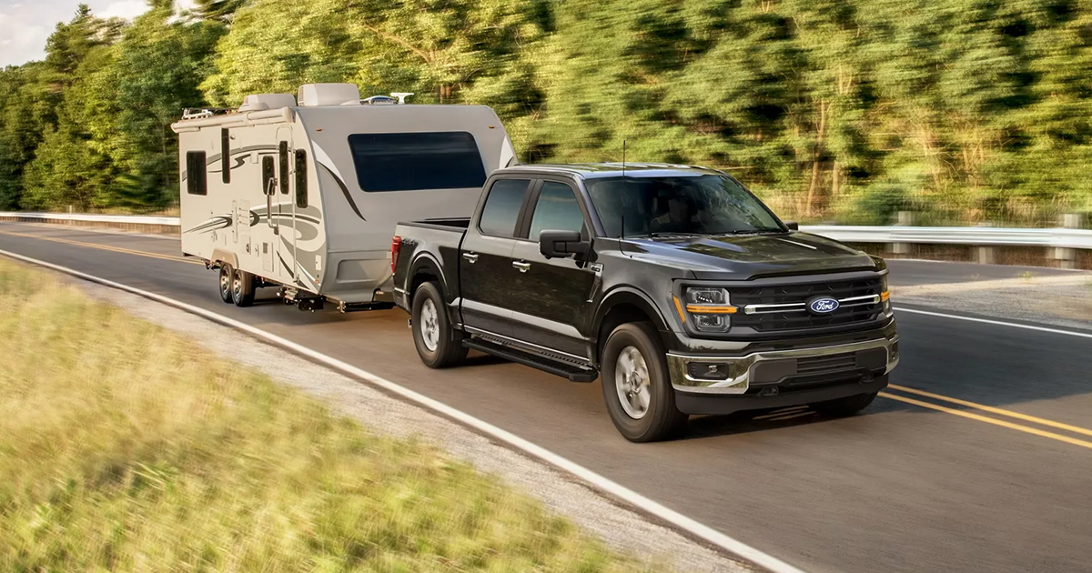 Black Ford F-150 pickup truck towing a white travel trailer on a scenic forest highway with blurred greenery
