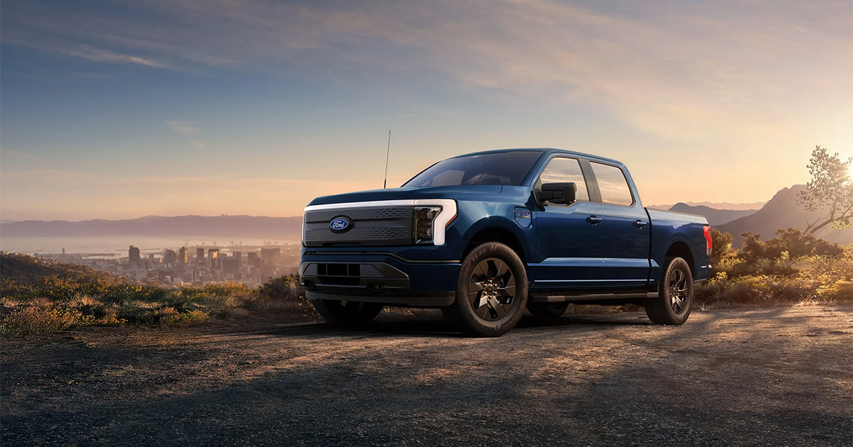 Blue Ford F-150 Lightning electric pickup truck parked on dirt road with city skyline and mountains at sunset