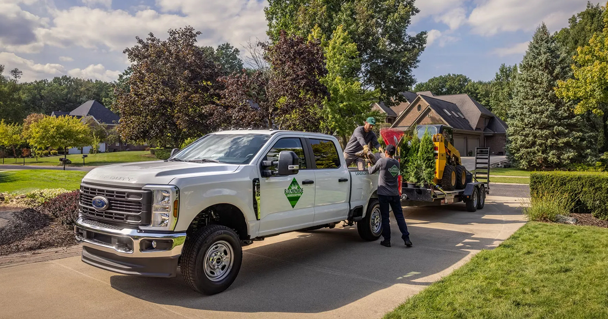 Ford F-250 work truck with landscaping trailer, two workers, and compact excavator in suburban neighborhood