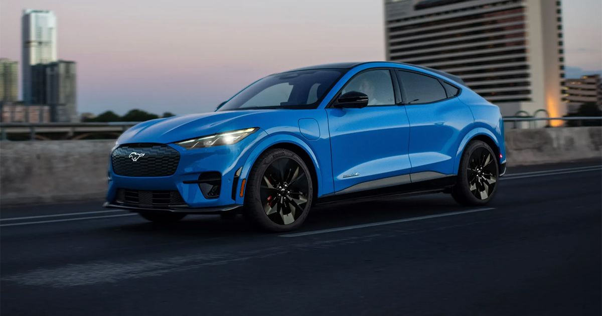 Blue Ford Mustang Mach-E electric SUV driving on urban road at sunset with city buildings in background
