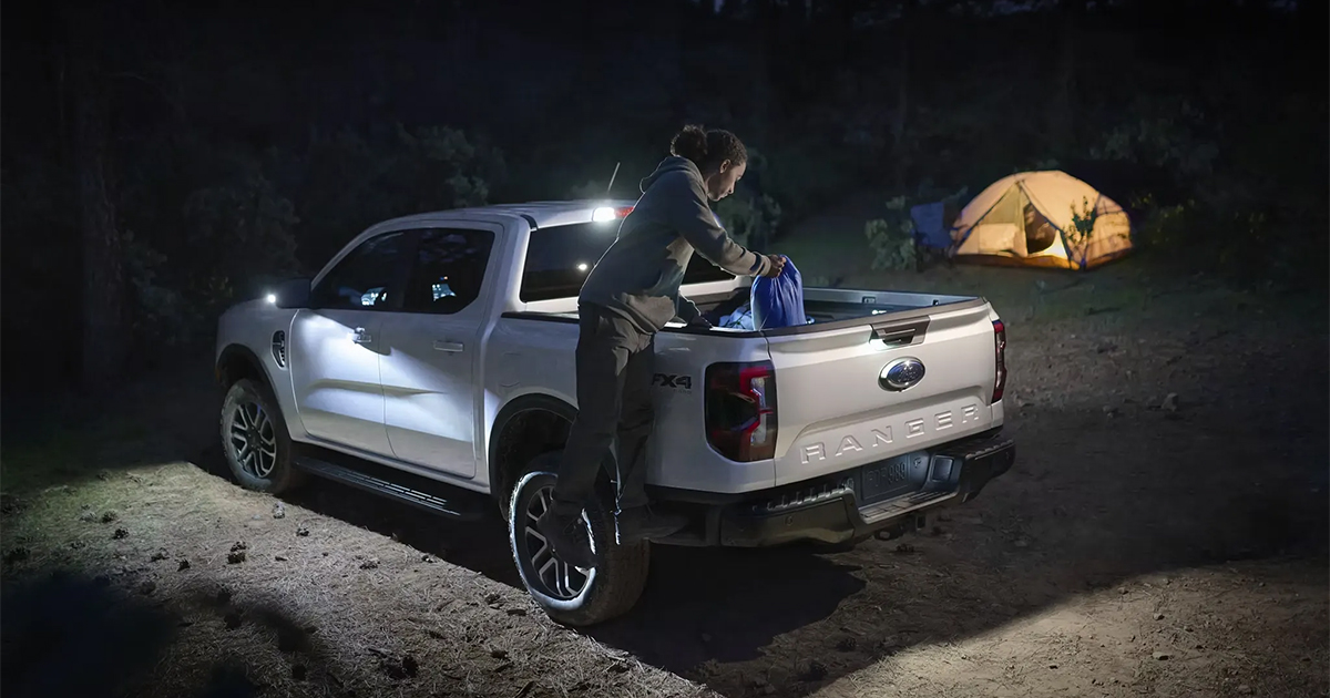 White Ford Ranger pickup truck at night with interior lighting, person loading gear near illuminated camping tent