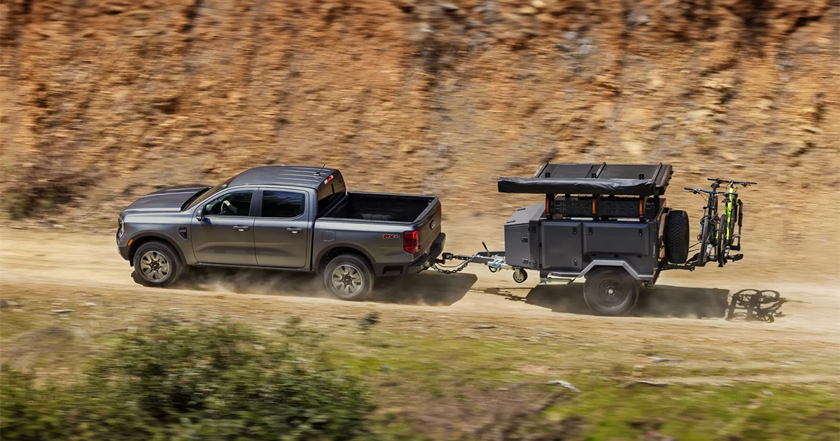 Gray Ford Maverick pickup truck towing an off-road trailer with bikes, driving on dusty dirt trail