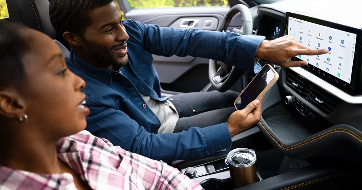 Two people in a Ford Explorer, driver using touchscreen infotainment system with smartphone, smiling