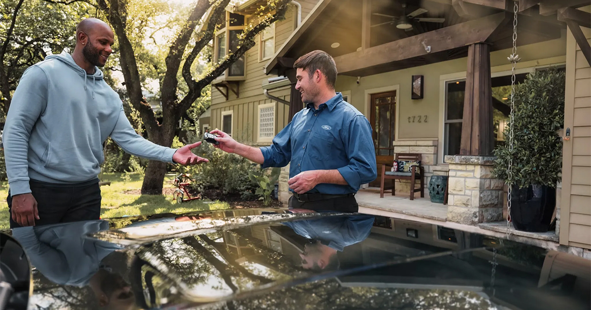 Two men exchanging car keys in front of a suburban home, with a Ford-branded blue shirt visible, reflecting car service or sale