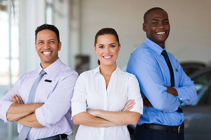 Professional automotive team of three smiling colleagues standing together in dealership, wearing business attire with confident poses