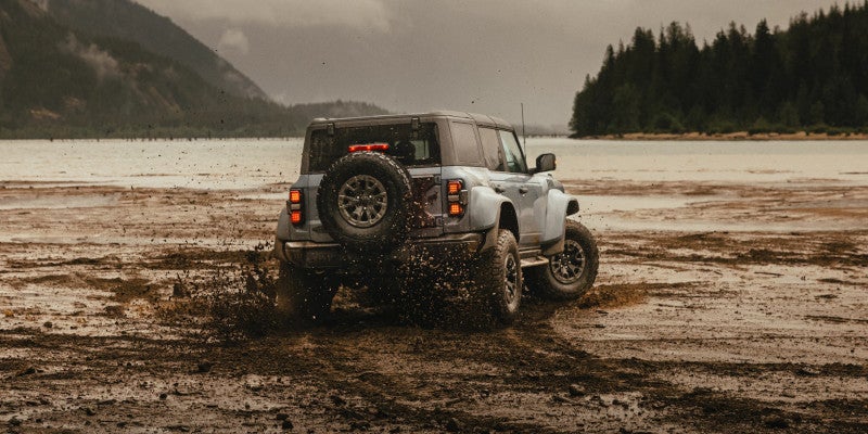 The Ford Bronco on rocky shore with mountains; two smaller Broncos off-roading