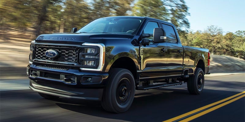 A black Ford Super Duty pickup truck driving fast on a paved road lined with trees.