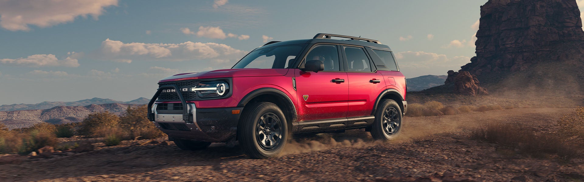 A bright pink/magenta Ford Bronco Sport drives across a rocky, desert landscape under a blue sky