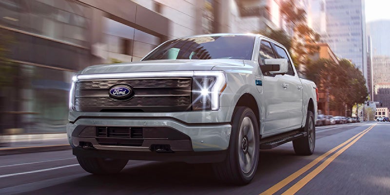 A light-colored Ford F-150 Lightning electric pickup truck driving on a bright city street.
