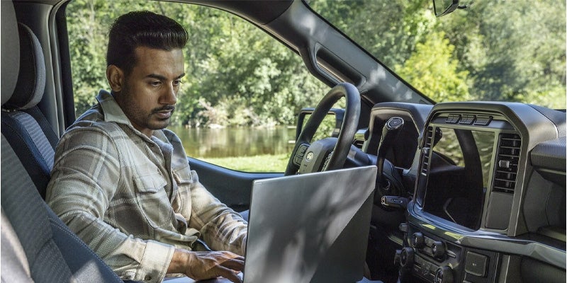 A man sitting in a Ford F-150 with the interior work surface open, working on a laptop near a lake.