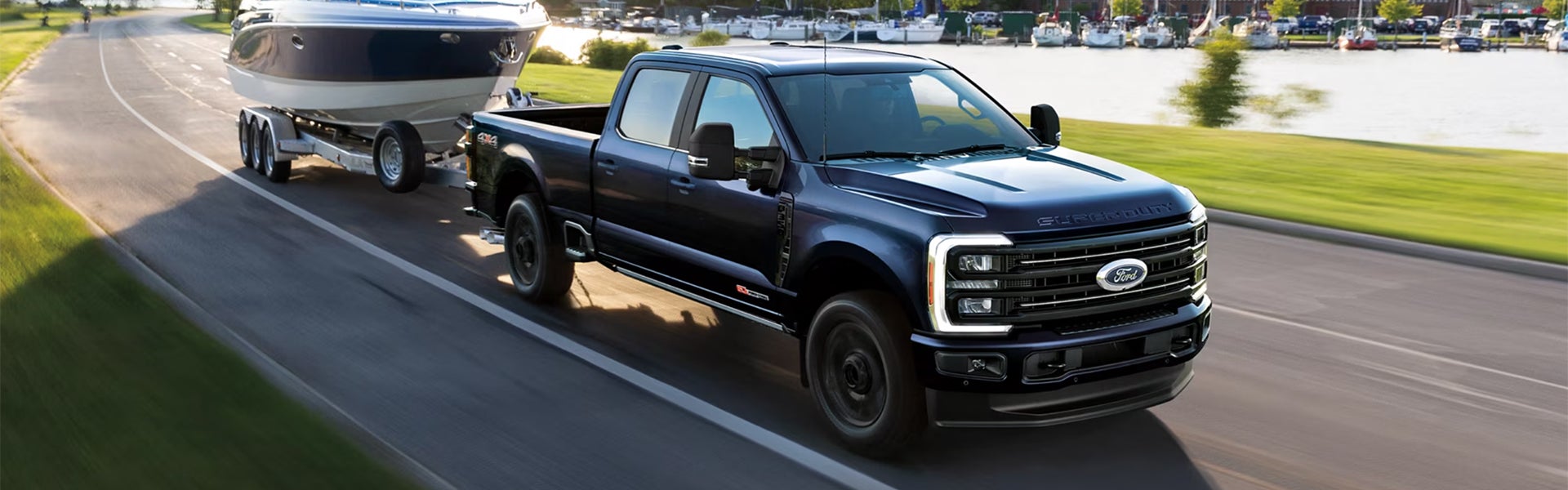 A dark blue Ford Super Duty truck towing a white and blue boat on a trailer, driving along a waterfront.